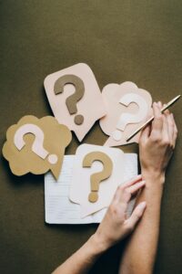 Hands holding pen next to paper question marks on notebook, symbolizing inquiry and creativity.