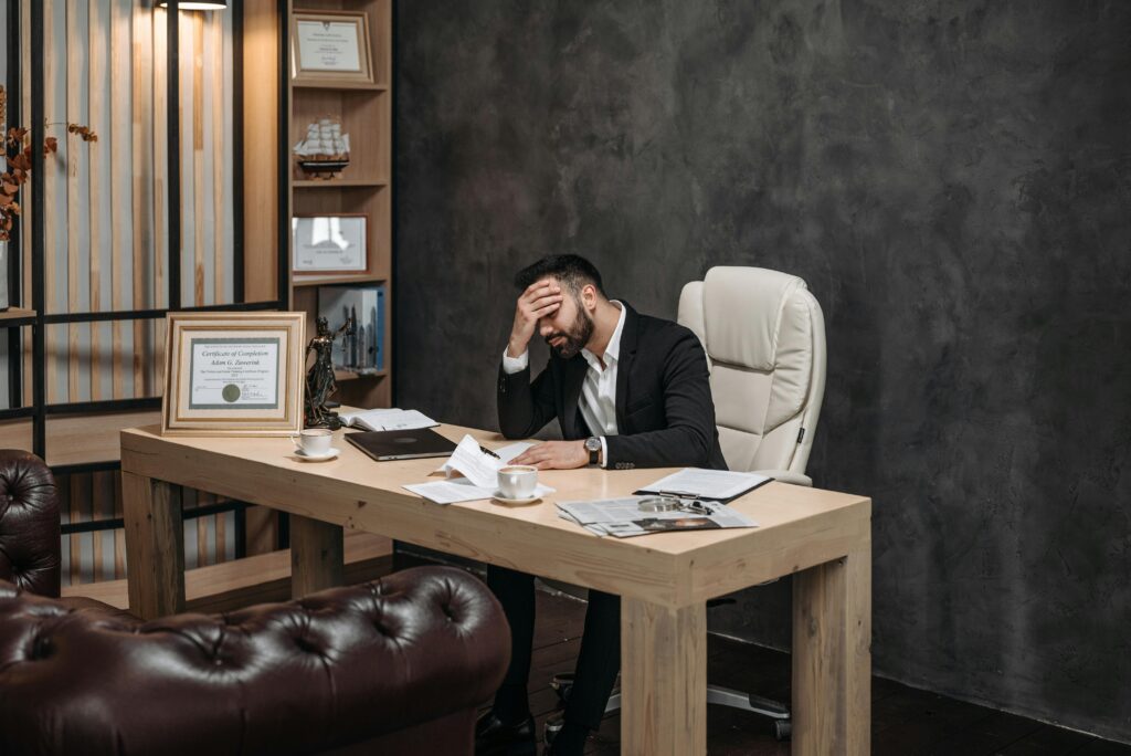 Businessman in a black suit looking stressed at an office desk, holding his head with papers scattered around.