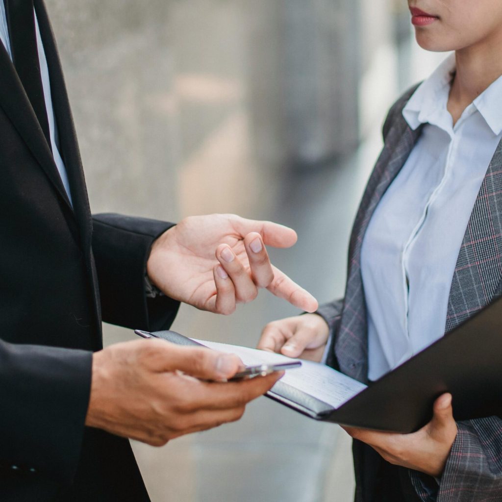 A businessman and businesswoman in formal attire engage in a focused discussion outdoors.